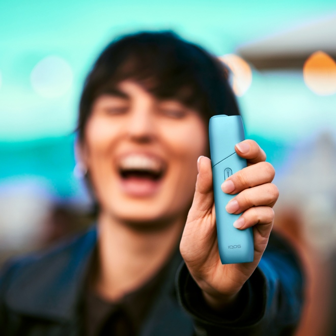 Hand holding a turquoise IQOS Originals One heated tobacco device with a guitar.